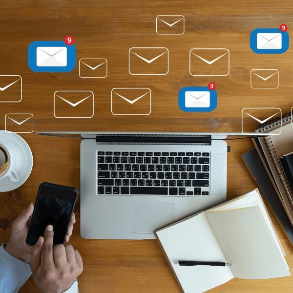 Person holding a phone at a desk with a laptop, coffee, notebooks, and floating envelope icons indicating new email notifications.