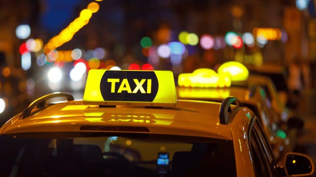 Yellow taxis lined up on a city street at night, with illuminated taxi signs and blurred lights in the background.