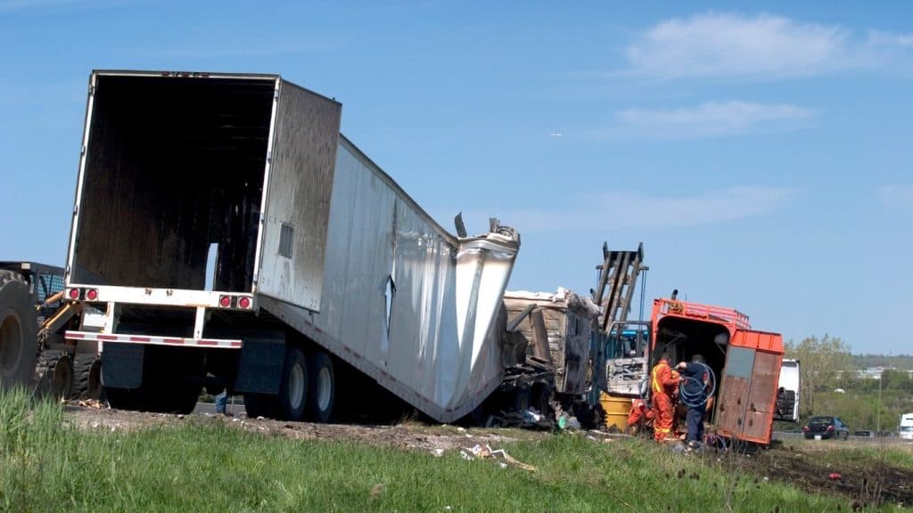 A semi-truck is overturned on the roadside, with a damaged trailer. People and machinery are assessing the scene under a clear sky.