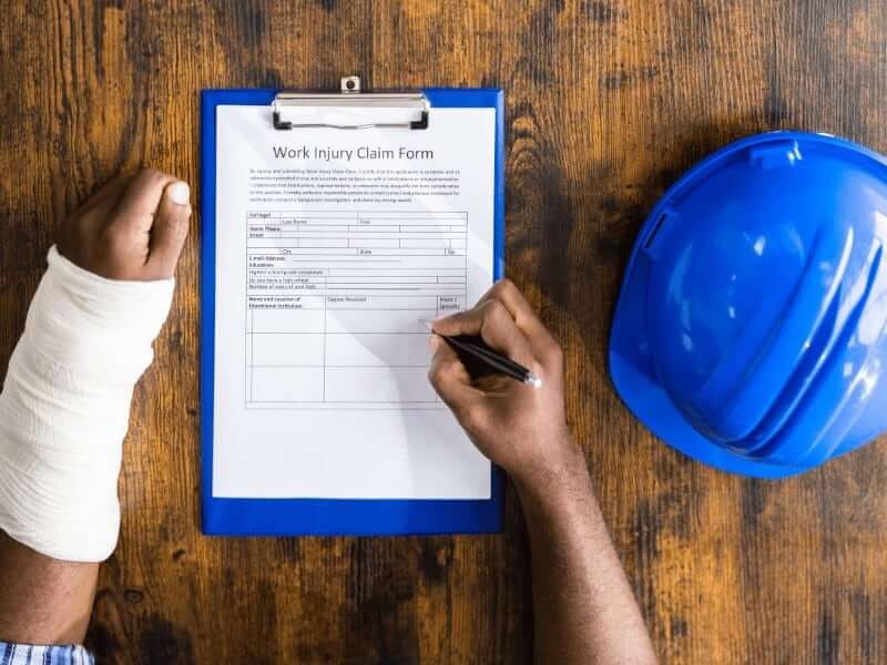 Person with a bandaged arm fills out a work injury claim form on a clipboard, with a blue hard hat on the wooden table.